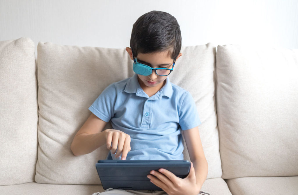 A boy is sitting on a sofa with a cover on hir right eye and is watching on his tablet with his lazy eye.