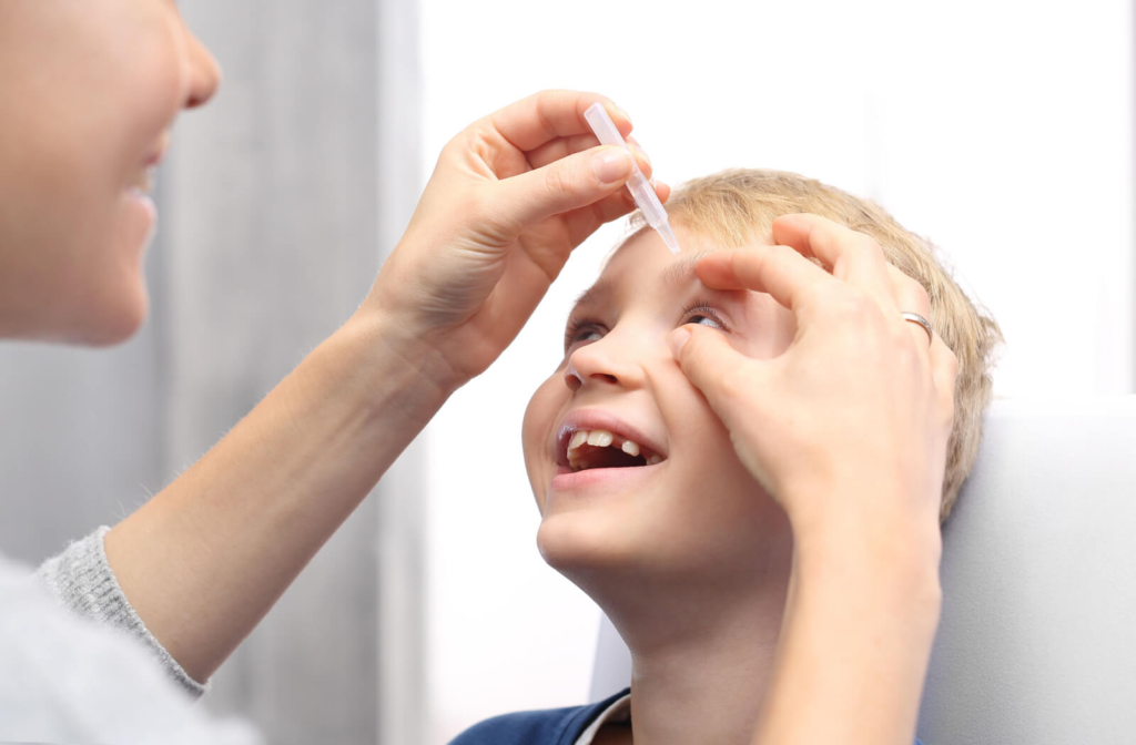 A young boy tilting his head back and smiling while a woman applies eye drops in his left eye.
