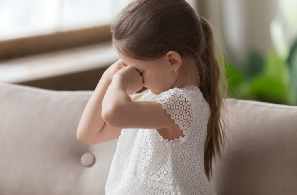 A young girl sitting on a couch and rubbing her eyes with both hands.