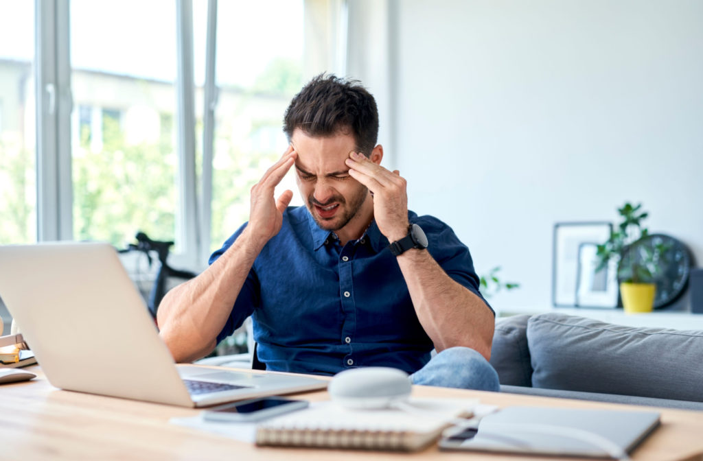 A man sitting at a desk with an open laptop and rubbing his temples because of a headache caused by an eye condition.