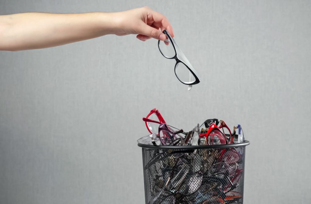 Woman's outstretched arm holding a pair of glasses over a waste basket full of eyeglasses.