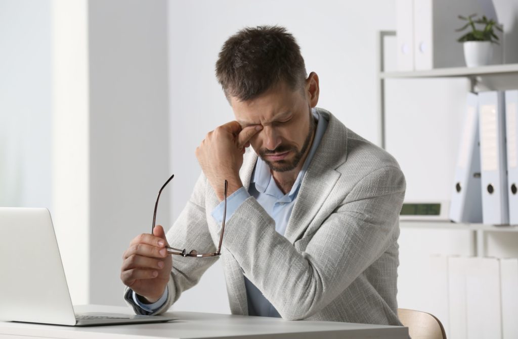 A man takes his glasses off and rubs his watery eyes after working on his laptop at home.