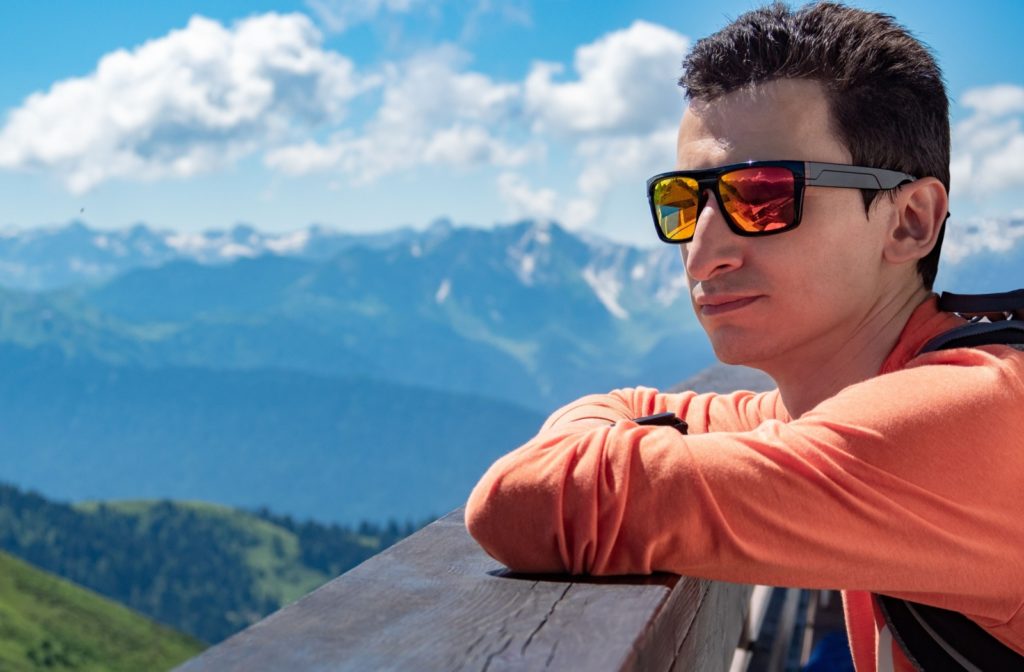 A man rests against a wooden railing, enjoying a beautiful view while wearing polarized sunglasses.