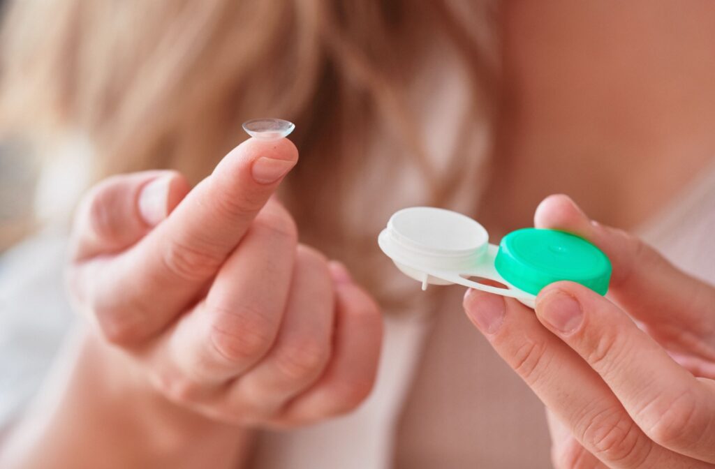 A person holds a contact lens on their fingertip with an open lens case in the other hand, preparing to insert or clean the lens.